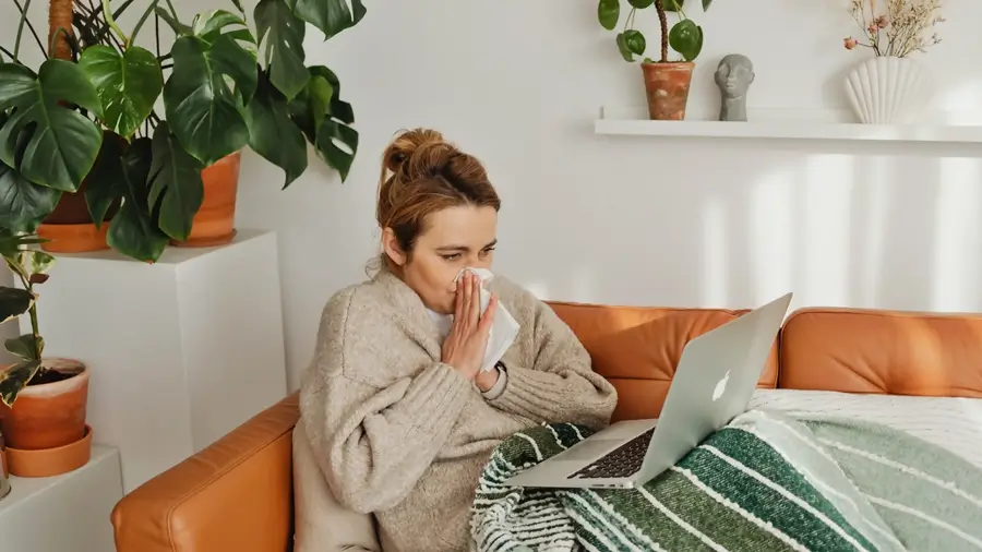 mujer sentada en un mueble sonandose la nariz mientras trabaja con su laptop