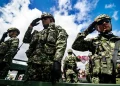 oldados con uniforme camuflado realizan saludo militar durante un acto público al aire libre, con cielo azul y banderas al fondo