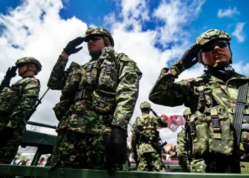 oldados con uniforme camuflado realizan saludo militar durante un acto público al aire libre, con cielo azul y banderas al fondo