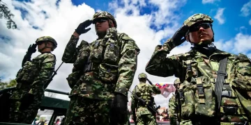 oldados con uniforme camuflado realizan saludo militar durante un acto público al aire libre, con cielo azul y banderas al fondo