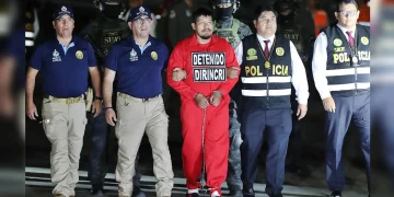 Detenido con uniforme rojo escoltado por agentes de INTERPOL y Policía Nacional del Perú durante operativo nocturno.