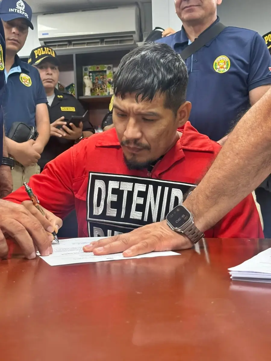 Detenido con cartel “DETENIDO” firmando documentos en oficina, rodeado por agentes de INTERPOL y Policía Nacional del Perú.