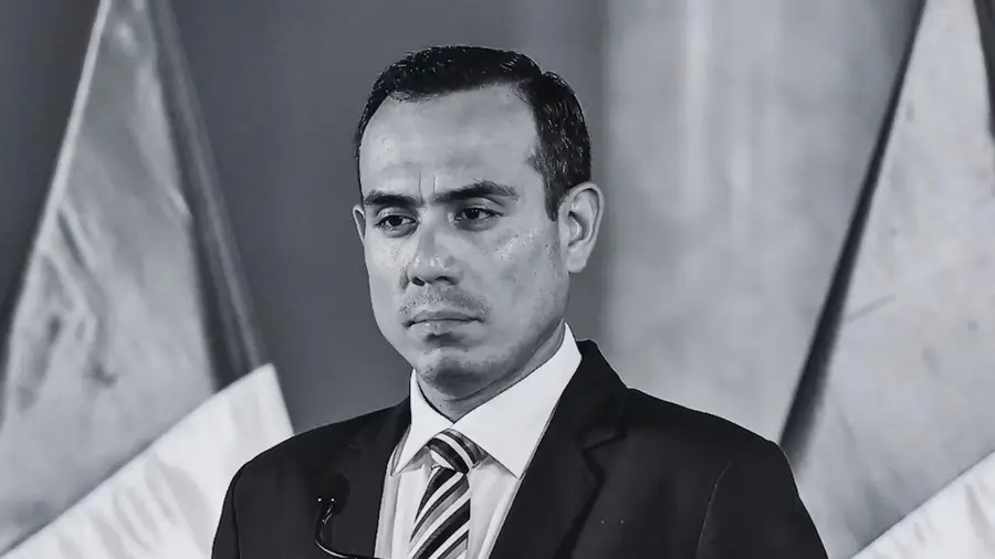 Hombre con traje y corbata, de expresión seria, en una imagen en blanco y negro, con banderas desenfocadas al fondo durante una declaración pública.