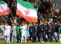 Jugadores de la selección de fútbol de Irán caminan por el campo tras un partido, mientras ondean banderas nacionales gigantes frente a una tribuna llena de espectadores.
