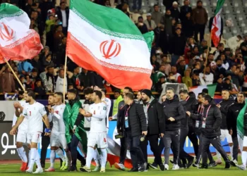 Jugadores de la selección de fútbol de Irán caminan por el campo tras un partido, mientras ondean banderas nacionales gigantes frente a una tribuna llena de espectadores.