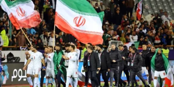 Jugadores de la selección de fútbol de Irán caminan por el campo tras un partido, mientras ondean banderas nacionales gigantes frente a una tribuna llena de espectadores.