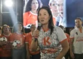 Keiko Fujimori hablando con micrófono en un mitin político; viste una camiseta blanca con el logo naranja de Fuerza Popular y de fondo se ve una pantalla gigante con su propia imagen.