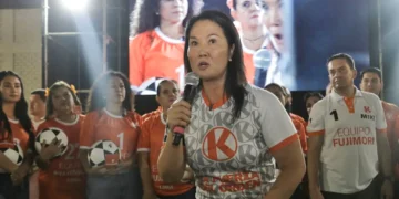 Keiko Fujimori hablando con micrófono en un mitin político; viste una camiseta blanca con el logo naranja de Fuerza Popular y de fondo se ve una pantalla gigante con su propia imagen.
