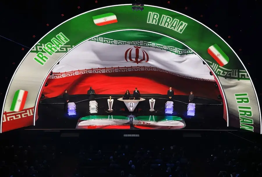 Escenario de sorteo oficial con una pantalla curva gigante que muestra la bandera de Irán y el texto "IR IRAN"; en el centro se observan presentadores, bombos de sorteo y el trofeo del Mundial.