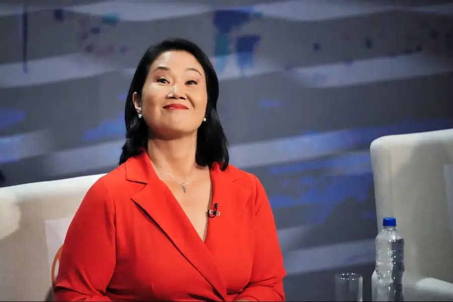 Keiko Fujimori sonriendo durante una entrevista o debate televisivo, vistiendo un saco rojo y un collar con una cruz, en un set con fondo azul y gris.